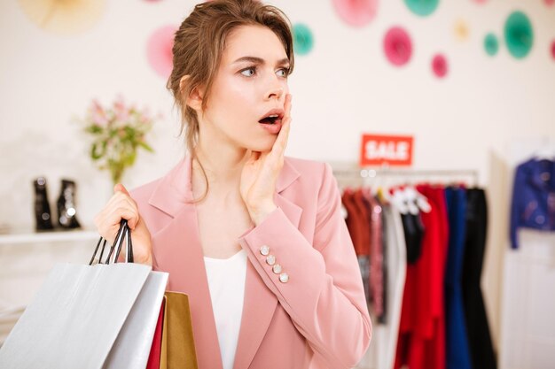 Chica joven en chaqueta rosa mirando a un lado asombrosamente con bolsas de compras en la mano en la tienda de ropa. Señora sorprendida de pie en la boutique con perchero de venta en el fondo