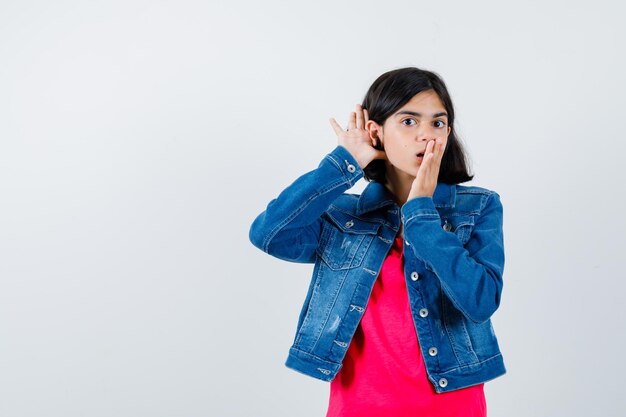 Chica joven cogidos de la mano cerca de la oreja para escuchar algo y cubriendo la boca con la mano en camiseta roja y chaqueta de jean y mirando sorprendido. vista frontal.