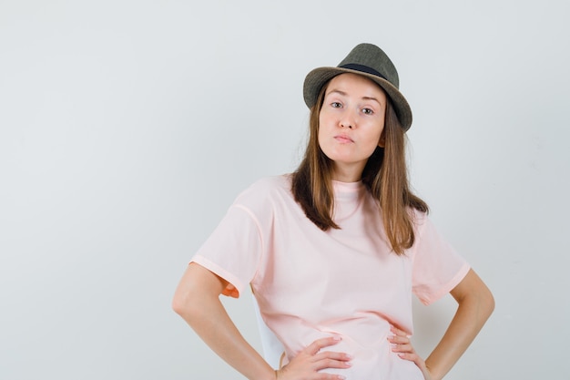 Chica joven cogidos de la mano en la cintura con camiseta rosa, sombrero y mirando disgustado, vista frontal.