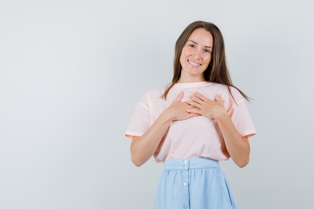 Foto gratuita chica joven cogidos de la mano en el corazón en camiseta, falda y mirando alegre. vista frontal.