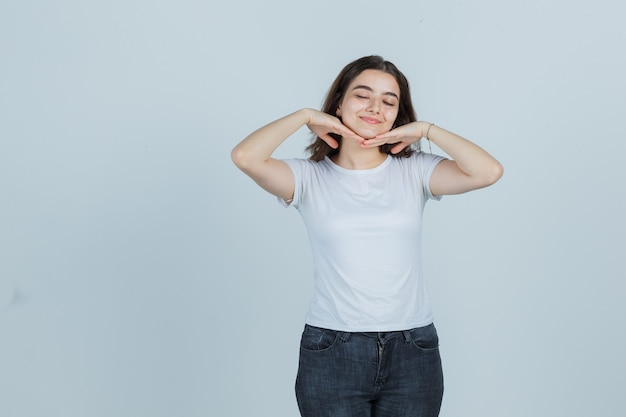 Chica joven cogidos de la mano debajo de la barbilla en camiseta, jeans y luciendo linda. vista frontal.