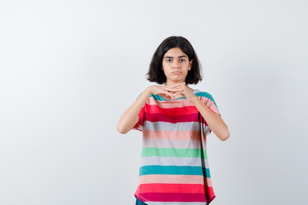 Chica joven cogidos de la mano debajo de la barbilla en camiseta a rayas de colores y mirando seria, vista frontal.