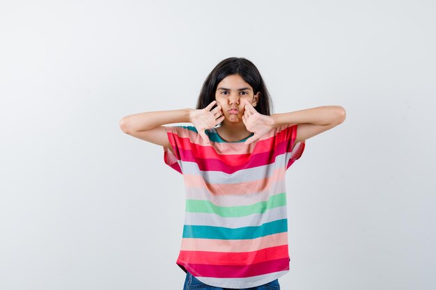 Chica joven cogidos de la mano en las mejillas en camiseta a rayas de colores y luciendo lindo. vista frontal.