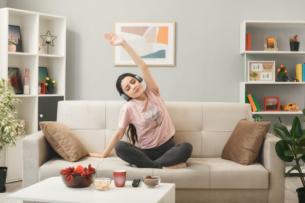 Chica joven complacida con audífonos haciendo yoga sentado en el sofá detrás de la mesa de café en la sala de estar