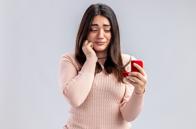 Chica joven complacida en el día de San Valentín sosteniendo y mirando el anillo de bodas aislado sobre fondo blanco.