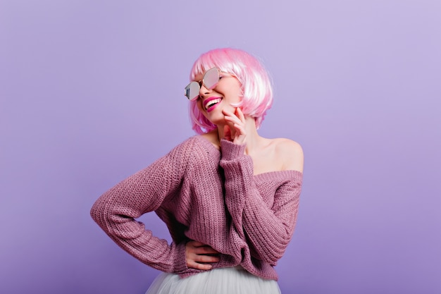 Chica joven complacida en peluca y gafas de sol divirtiéndose Foto de modelo de mujer maravillosa con cabello rosado sonriendo mientras baila en la pared púrpura.