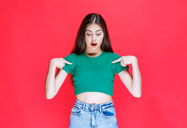 Chica joven confiada de pie y apuntando hacia abajo sobre fondo rojo.