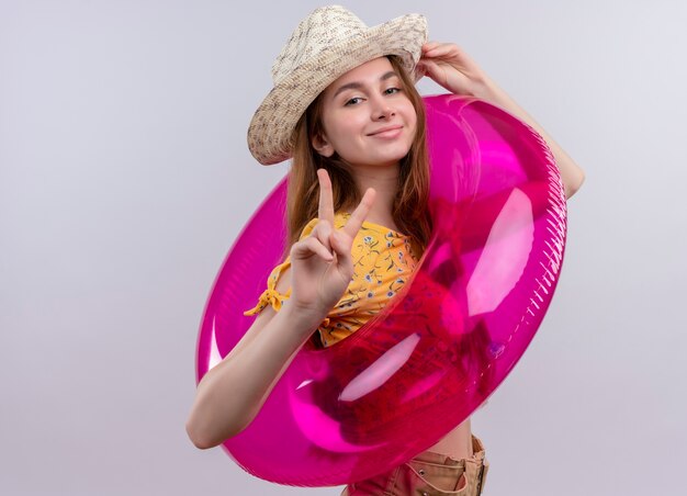 Chica joven confiada con sombrero y anillo de natación haciendo el signo de la paz y poniendo la mano en el sombrero en un espacio en blanco aislado con espacio de copia