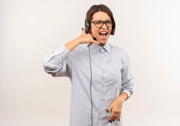 Chica joven confidente del centro de llamadas con gafas y auriculares haciendo distintivo de llamada aislado en blanco con espacio de copia
