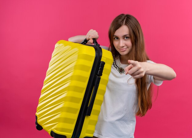 Chica joven confidente que viaja levantando la maleta y señalando el espacio rosa aislado