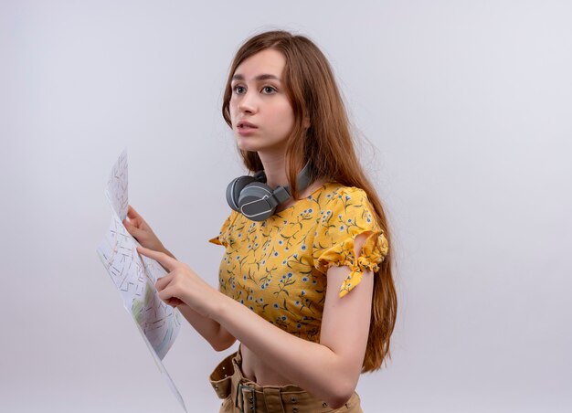 Chica joven confundida con auriculares en el cuello y sosteniendo el mapa y apuntando a él en un espacio en blanco aislado con espacio de copia