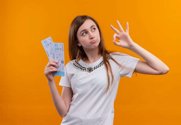 Chica joven confusa que viaja con billetes de avión haciendo bien firmar en el espacio naranja aislado