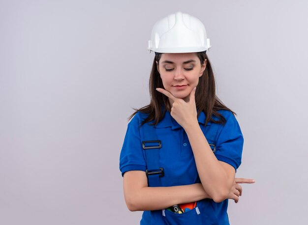 Chica joven constructor con casco de seguridad blanco y uniforme azul pone la mano en la barbilla y mira hacia abajo sobre fondo blanco aislado con espacio de copia