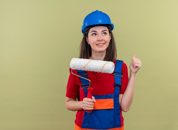 Chica joven constructora complacida con casco de seguridad azul sostiene el rodillo de pintura y levanta el puño sobre fondo verde aislado con espacio de copia