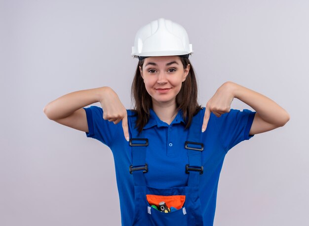 Chica joven constructora complacida con casco de seguridad blanco y uniforme azul apuntando hacia abajo con ambas manos sobre fondo blanco aislado