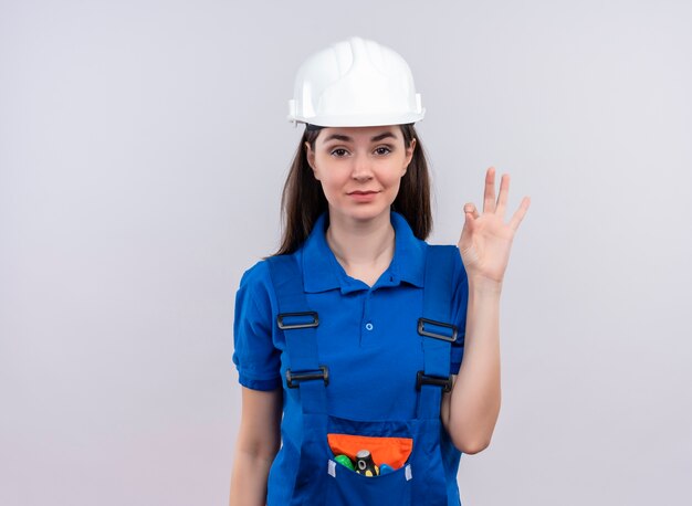 Chica joven constructora confiada con casco de seguridad blanco y gestos uniformes azules bien sobre fondo blanco aislado con espacio de copia