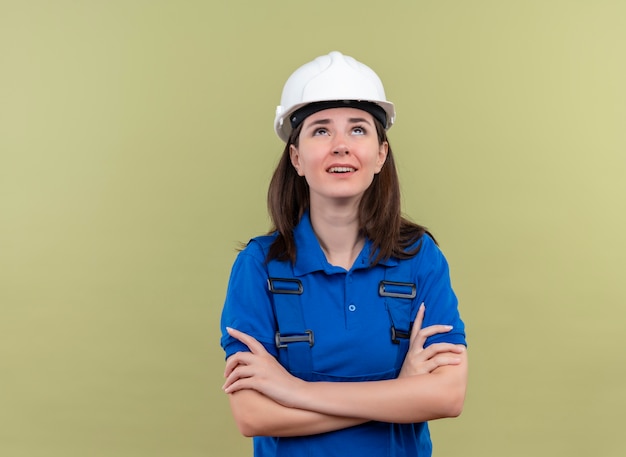 Chica joven constructora molesta con casco de seguridad blanco y uniforme azul cruzó los brazos y mira hacia arriba sobre fondo verde aislado con espacio de copia