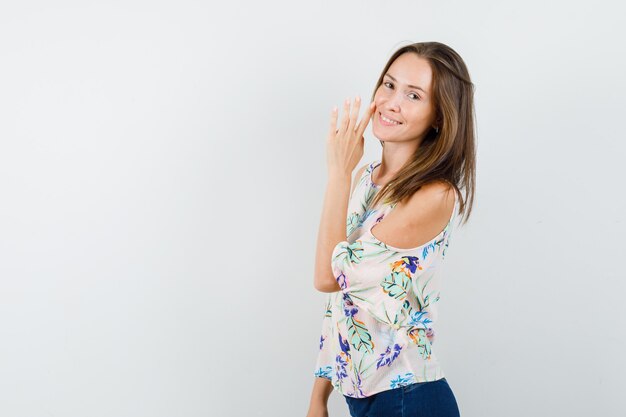 Chica joven contando secretos detrás de la mano en camisa, jeans y mirando alegre.