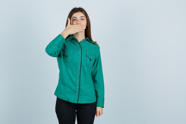 Chica joven cubriendo la boca con la mano en blusa verde, pantalón negro y mirando avergonzado, vista frontal.