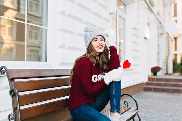 Chica joven de cuerpo entero con pelo largo con gorro de punto, jeans y guantes blancos sentado en un banco en la calle. Ella tiene un corazón rojo caramelo, sonriendo.