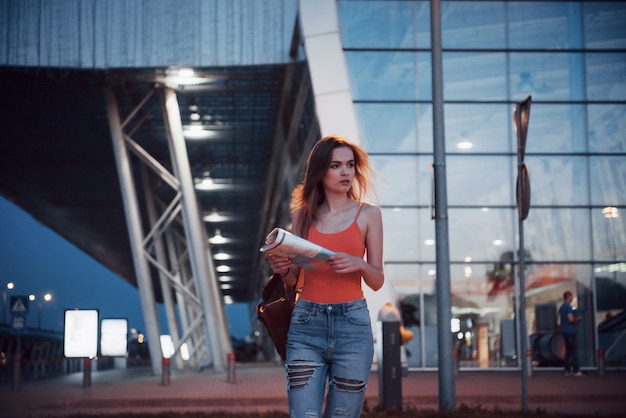 La chica joven cuesta por la noche cerca de la terminal del aeropuerto o estación y lee el mapa de la ciudad y busca el hotel. Lindo turista con mochilas determina el concepto de viaje