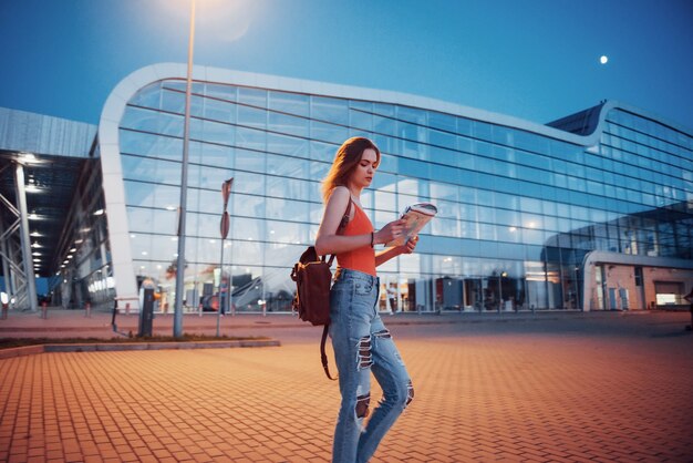 La chica joven cuesta por la noche cerca de la terminal del aeropuerto o estación y lee el mapa de la ciudad y busca el hotel. Lindo turista con mochilas determina el concepto de viaje