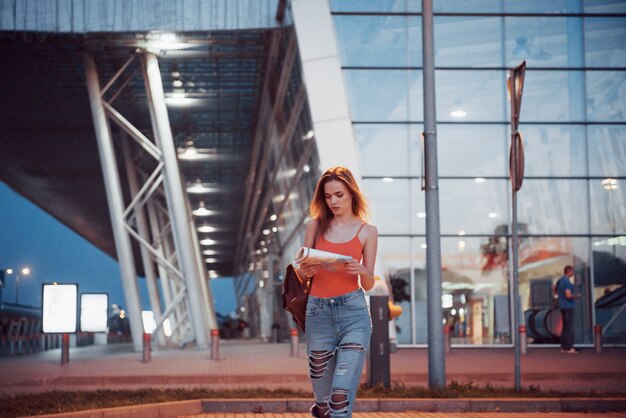 La chica joven cuesta por la noche cerca de la terminal del aeropuerto o estación y lee el mapa de la ciudad y busca el hotel.