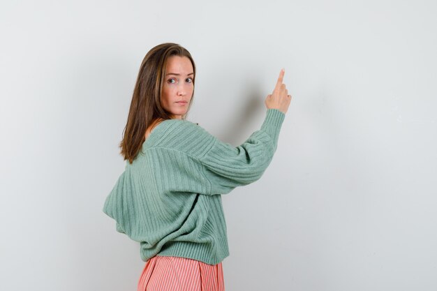 Foto gratuita chica joven dando marcha atrás, apuntando a la pared con el dedo índice, mirando a la cámara en prendas de punto, falda y mirando seductor, vista frontal.