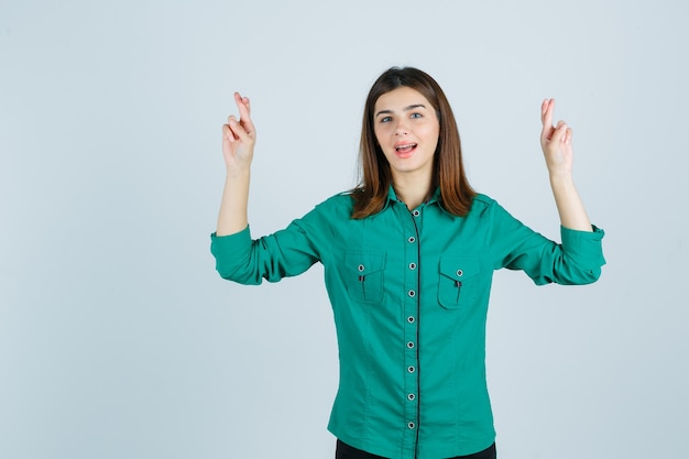 Chica joven con los dedos cruzados, manteniendo la boca abierta en blusa verde, pantalón negro y luciendo feliz. vista frontal.