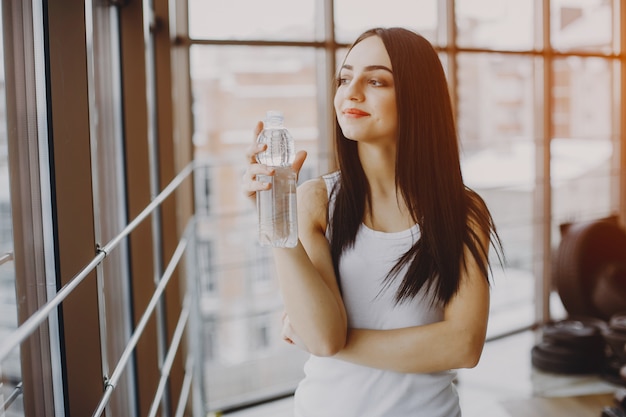 chica joven y delgada con una camisa blanca y polainas grises de pie en un gimnasio con botella