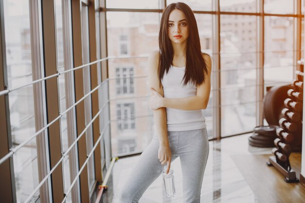 chica joven y delgada con una camisa blanca y polainas grises de pie en un gimnasio con botella