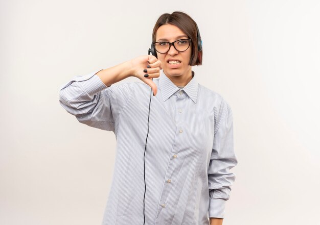 Chica joven descontenta del centro de llamadas con gafas y auriculares mostrando el pulgar hacia abajo aislado sobre fondo blanco con espacio de copia