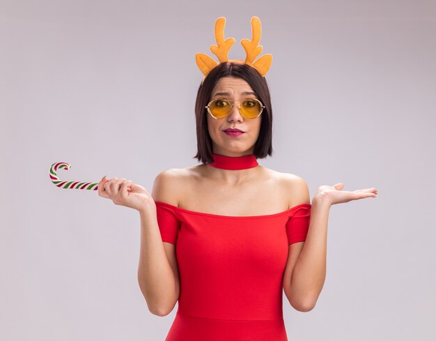 Chica joven despistada con diadema de cuernos de reno y gafas sosteniendo bastón de caramelo de Navidad de Navidad mostrando la mano vacía mirando a cámara aislada sobre fondo blanco