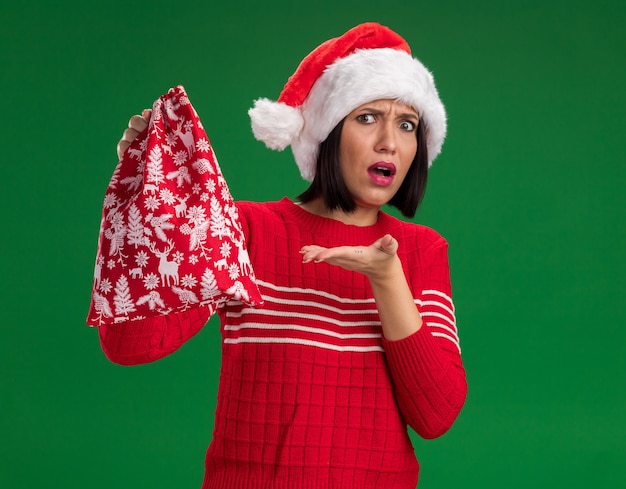 Chica joven despistada con gorro de Papá Noel sosteniendo y apuntando al saco de regalo de Navidad aislado en la pared verde