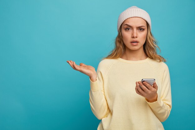 Chica joven despistada con sombrero de invierno sosteniendo teléfono móvil mostrando la mano vacía