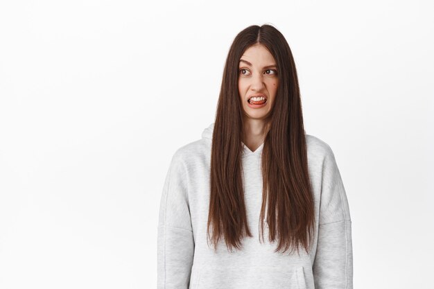 Chica joven divertida con cabello largo, mirada disgustada a un lado, mirando el contenido encogido, luciendo mal producto horrible con aversión, mostrando la lengua y haciendo muecas, de pie sobre una pared blanca