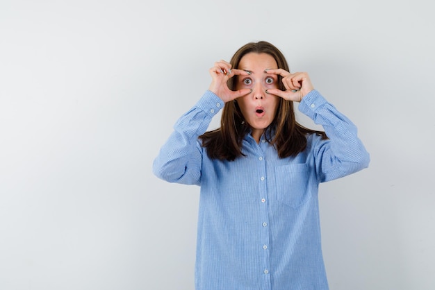 La chica joven divertida está abriendo los ojos con los dedos en el fondo blanco