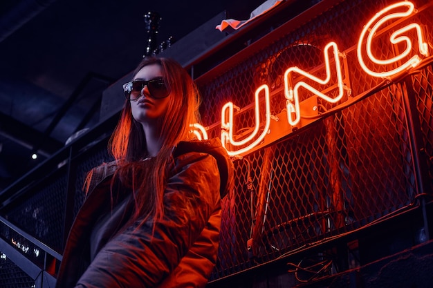 Chica joven elegante con un abrigo con capucha y gafas de sol de pie en las escaleras en la discoteca subterránea con interior industrial