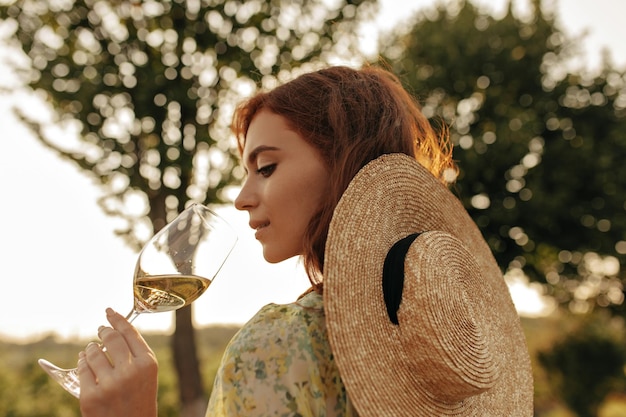 Chica joven elegante con peinado astuto en sombrero de paja moderno y traje de verano fresco posando con copa con champán al aire libre