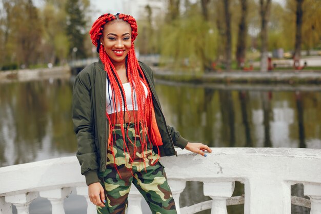 Una chica joven y elegante de piel oscura con rastas rojas caminando en el parque de verano cerca del río