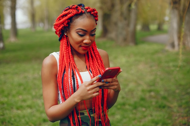 Una chica joven y elegante de piel oscura con rastas rojas caminando en el parque de verano con teléfono