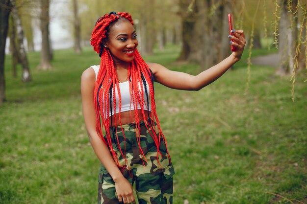 Una chica joven y elegante de piel oscura con rastas rojas caminando en el parque de verano con teléfono