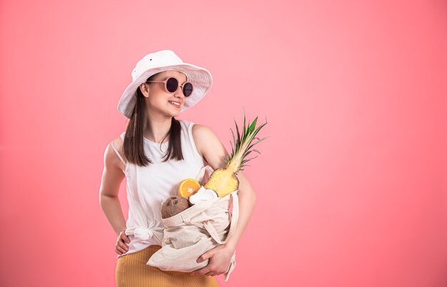 Chica joven elegante con sombrero blanco y gafas de sol sonríe y sostiene una bolsa ecológica con frutas exóticas en un espacio de copia de fondo rosa.