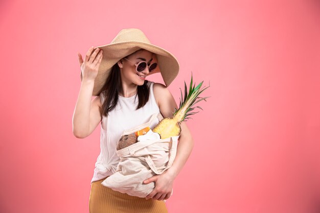 Chica joven elegante con sombrero grande y gafas de sol sonríe y sostiene una bolsa ecológica con frutas exóticas en un espacio de copia de fondo rosa.