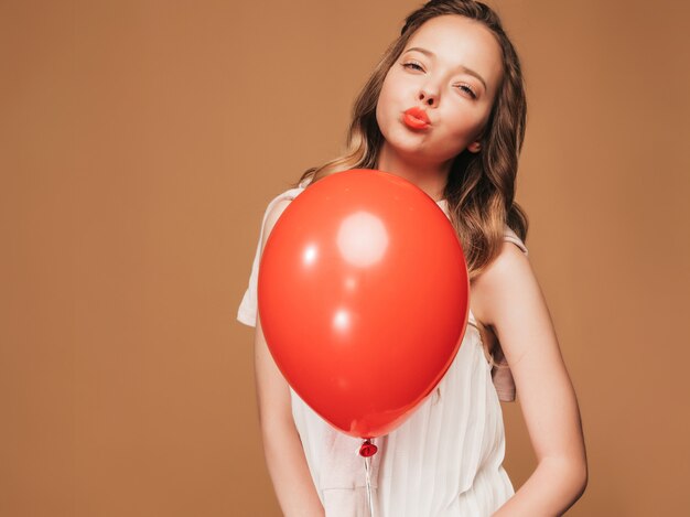 Chica joven emocionada que presenta en el vestido blanco del verano de moda. Modelo de mujer con globo rojo posando. Listo para fiesta y beso