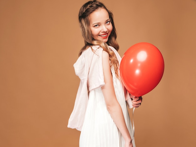 Chica joven emocionada que presenta en el vestido blanco del verano de moda. Modelo de mujer con globo rojo posando. Listo para la fiesta