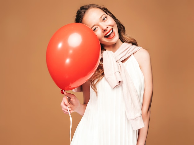 Chica joven emocionada que presenta en el vestido blanco del verano de moda. Modelo de mujer con globo rojo posando. Listo para la fiesta