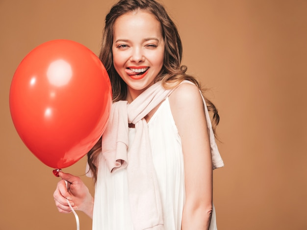 Chica joven emocionada que presenta en el vestido blanco del verano de moda. Modelo de mujer con globo rojo posando. Mostrando su lengua y lista para la fiesta