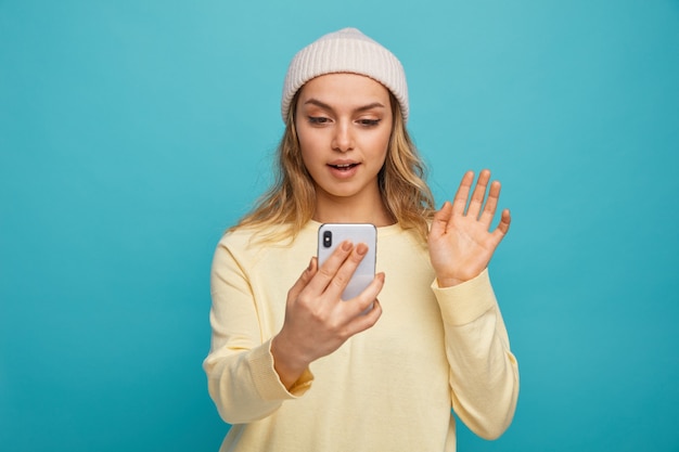 Chica joven emocionada con sombrero de invierno sosteniendo y haciendo gesto de hola