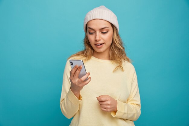 Chica joven emocionada con sombrero de invierno sosteniendo y mirando el teléfono móvil